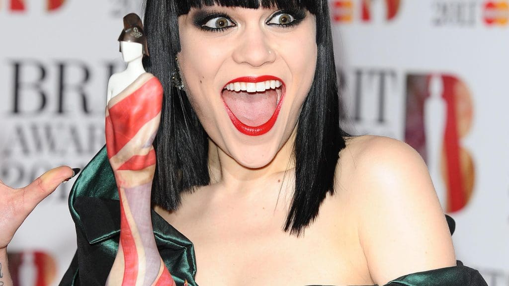 Image of Jessie J holding the Vivianne Westwood Brit Award Trophy. It's a close crop where she's looking delighted and posing open-mouthed wearing bright red lipstick.