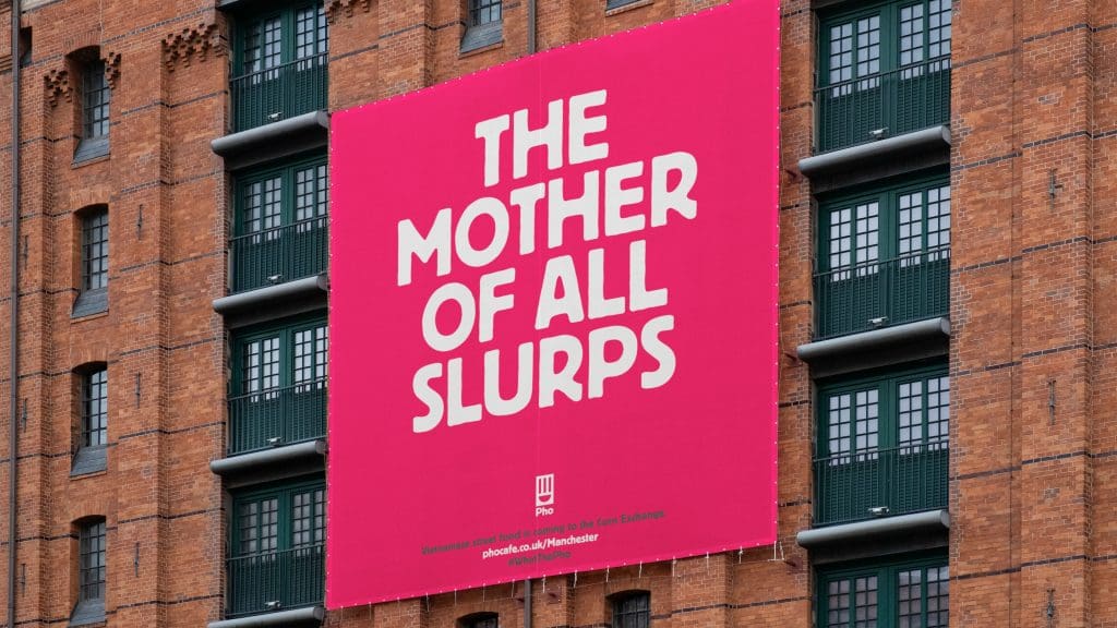 Photograph of a large brick-built warehouse-style building with a large hanging outdoor ad banner. The banner is red holding a Pho ad which says 'Mother of all slurps'.