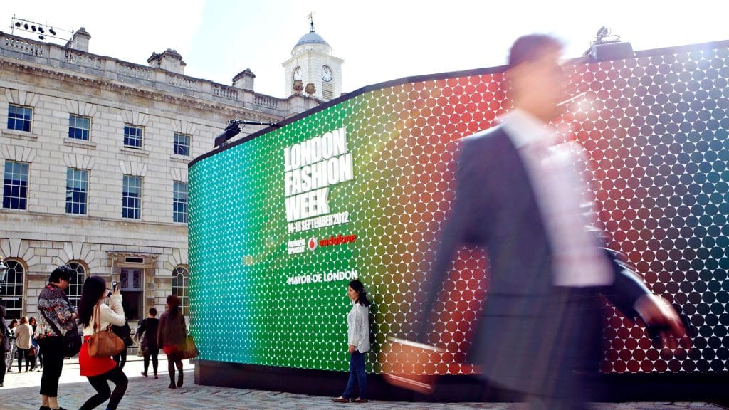 Large hoarding at London Fashion Week, uses the pattern of Jonathan Saunders to create a photographic backdrop for visitors. People are posing for photographs in front of the structure. 