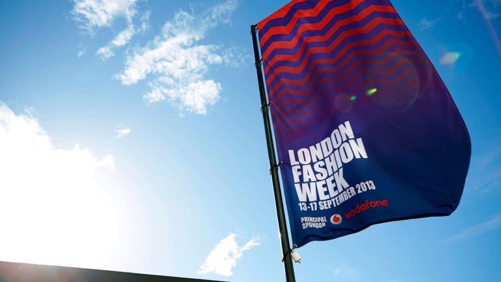 Large banner flag against a bright blue sky. The flag has the navy and red zigzag motive of Nicholas Kirkwood, and shows the onsite branding of London Fashion Week.