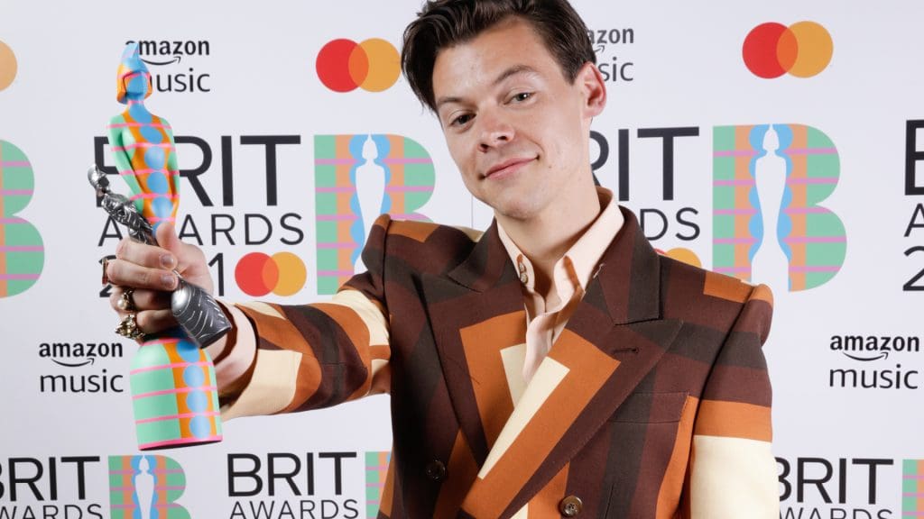 Image of Harry Styles dressed in a 60s style suit, holding the Yinka Ilori Brit Award trophy at arm's length, looking pleased.