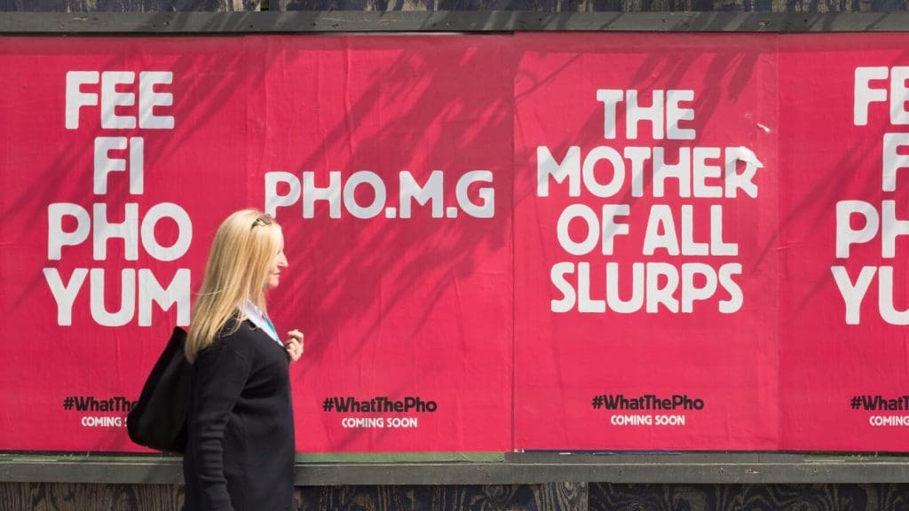 Photograph of a close-up woman walking past a flyposters with Pho ads using bright red backgrounds and white text. These read 'Fee Fi Pho Yum', 'Pho.m.g', and 'The mother of all slurps'.