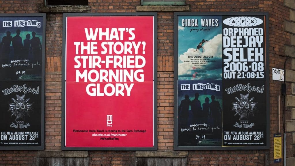 Photograph of a brick-built building with 3 fly-posting sites. The middle poster is a red Pho ad that says, 'What's the story? Stir-fried morning glory'.